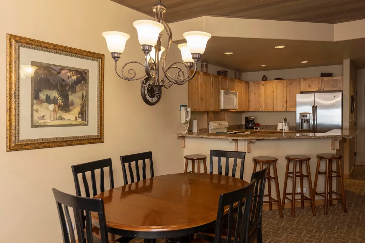 A welcoming dining area anchors the open living space, with a large wooden table set beneath a stylish chandelier and surrounded by comfortable chairs. The scene conveys a sense of community, ideal for family meals, celebrations, or casual gatherings. Artistic touches and the nearby kitchen counter hint at an easy flow between social spaces.