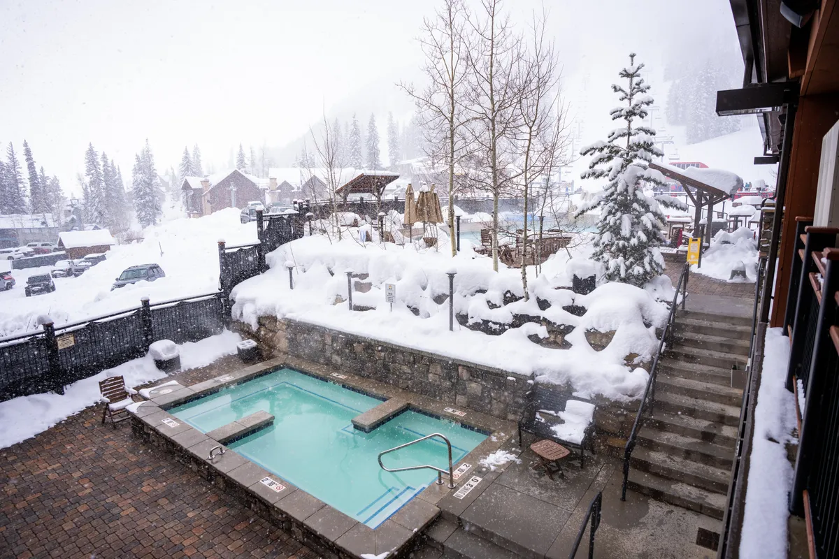 A cozy outdoor hot tub steams in the foreground, surrounded by gently falling snow and snow-laden trees, with stone steps leading to the resort grounds. The scene radiates relaxation after a day on the slopes.