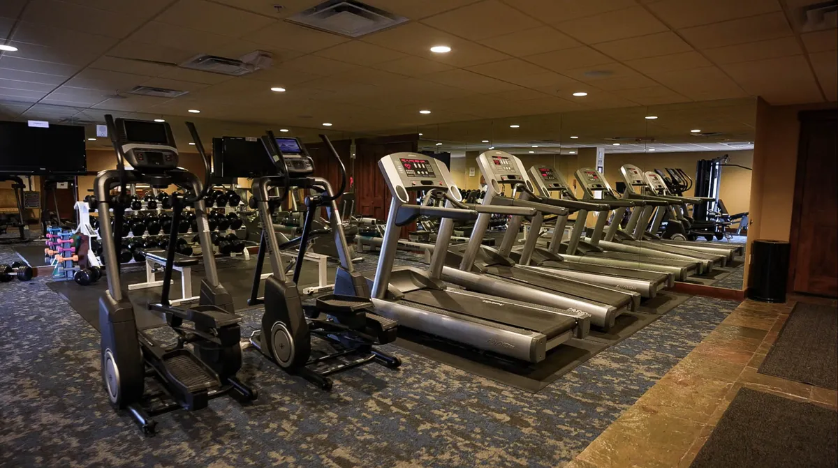 A vibrant, well-equipped fitness area featuring treadmills, ellipticals, dumbbells, a mirrored wall, and exercise mats. The atmosphere is energetic, supporting both solo and group workouts.