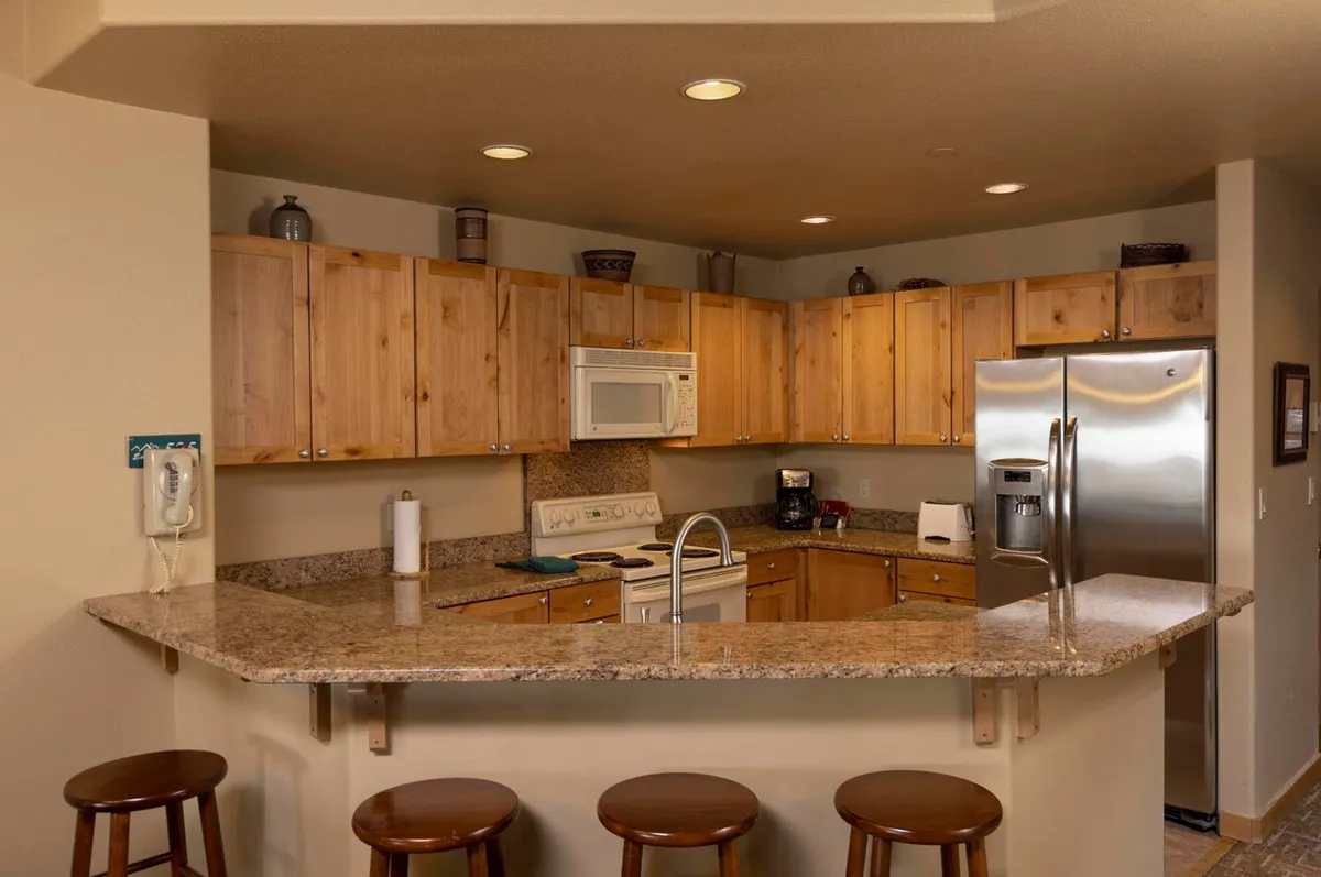 A warm, welcoming kitchen space featuring sleek granite countertops, rich wooden cabinets, and state-of-the-art stainless steel appliances. Bar stools line the counter, inviting guests to gather and participate in meal preparation or enjoy casual snacks. The mood is lively and communal, with subtle hints of activity like cooking or hosting family meals.