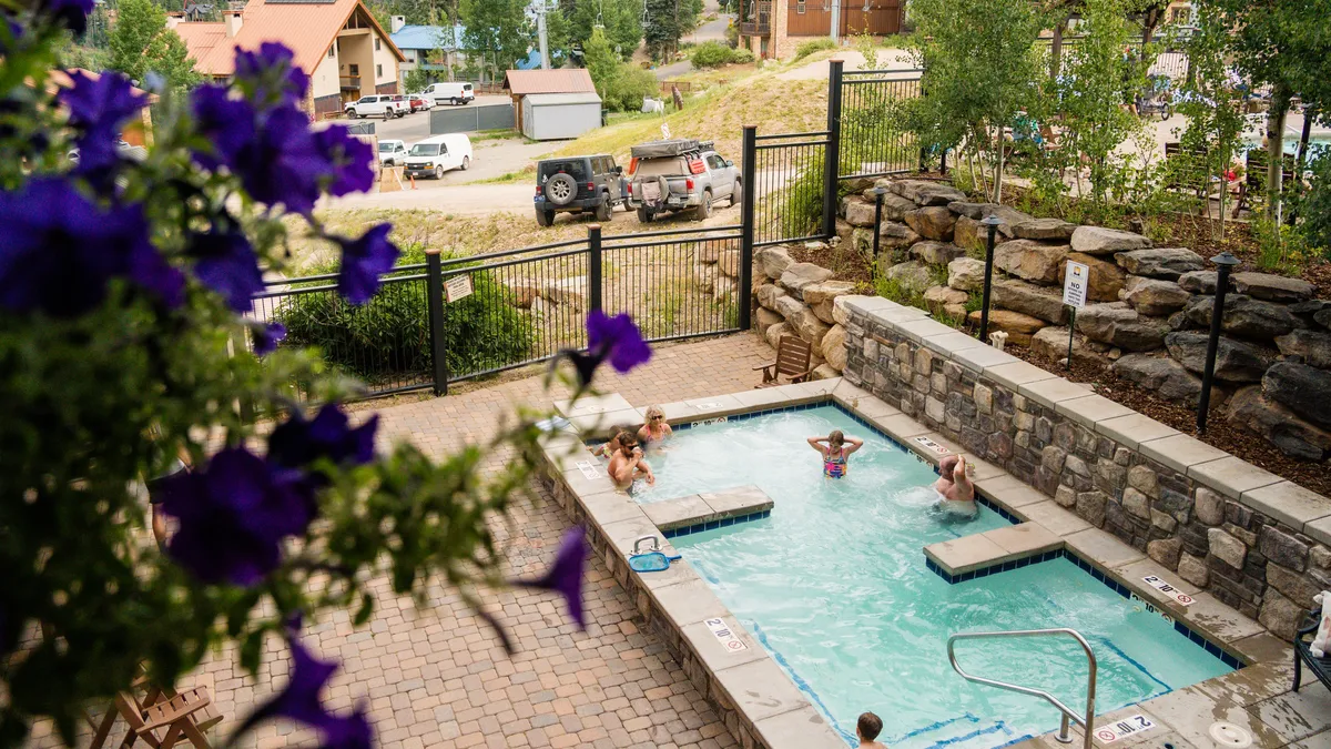 An inviting outdoor area with a hot tub surrounded by flowers, adjacent to a pool area with a stone wall and fence. The space is perfect for relaxation, socializing, and enjoying the scenery after a day of mountain activities.