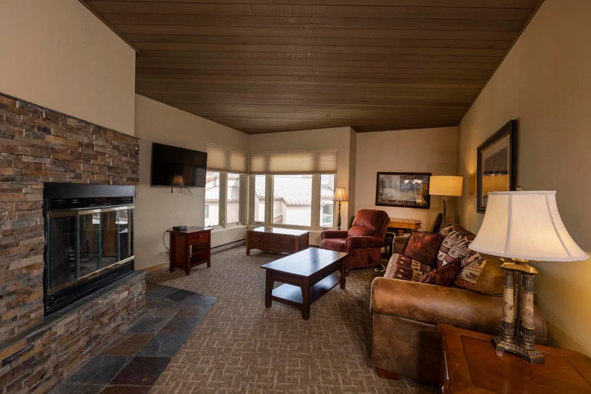 The image showcases a cozy seating area centered around a glowing fireplace, bathed in natural light from expansive windows that frame breathtaking mountain views. Plush seating invites guests to relax, whether it's curling up with a book, chatting with friends, or simply soaking in the tranquil alpine atmosphere.