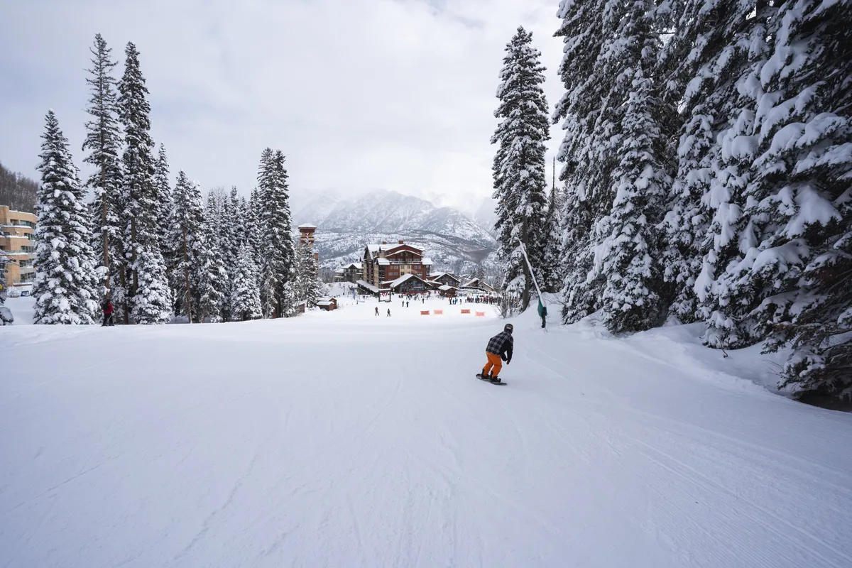 An exhilarating winter scene showcasing a snowboarder soaring down a snow-covered mountainside, with a ski resort lodge nestled among pine trees in the background. The atmosphere is energetic, embodying the winter fun and memorable moments unique to resort life.