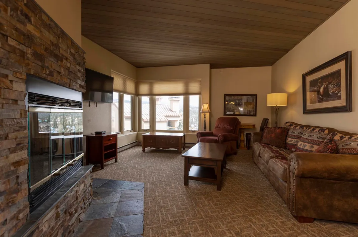 This inviting living room features a prominent fireplace surrounded by cozy seating, including a sofa and armchairs, all oriented to take full advantage of expansive windows that offer sweeping views of the Needles Mountains and ski slopes beyond. The space is bright and airy, perfect for both relaxing and socializing, with sunlight streaming in during the day and a warm glow from the fire in the evening.