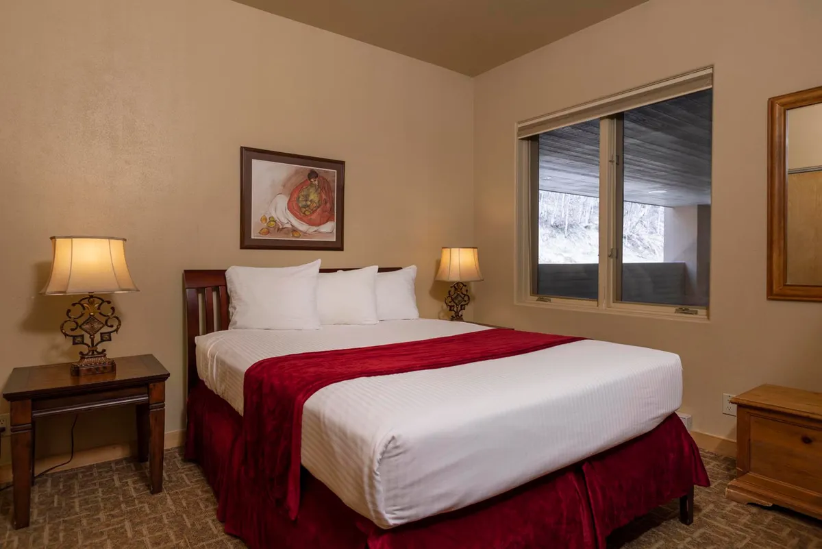 A bright, inviting bedroom featuring a plush bed with a deep red blanket, decorative lamps on each side, tasteful wall art, and a large window that fills the space with natural sunlight. The overall mood is warm and restful, perfect for relaxation and uninterrupted sleep.