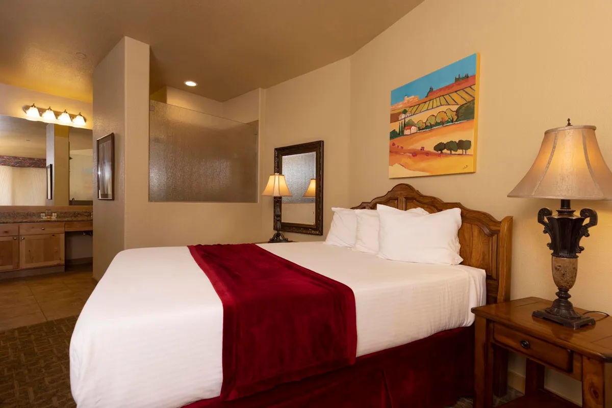A welcoming bedroom scene featuring a plush bed adorned with a vibrant red throw, complemented by soft natural light streaming in. Local artwork graces the walls, and a bedside lamp rests on a nightstand, creating an intimate and restful atmosphere.